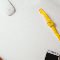 hands typing on a white keyboard. A yellow wristwatch lies on a white table nearby.