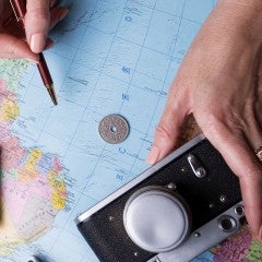 Hands hold a telephone and a compass on a table topped by a map. A croissant and a cup of coffee are on the table nearby.