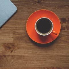 A cup of coffee on an orange saucer sits on a wooden desk. A laptop is seen in the upper corner of the screen.