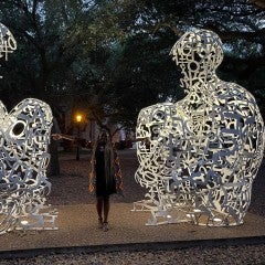two white sculptures of humans seated in front of each other, with a woman in the middle with her arms open